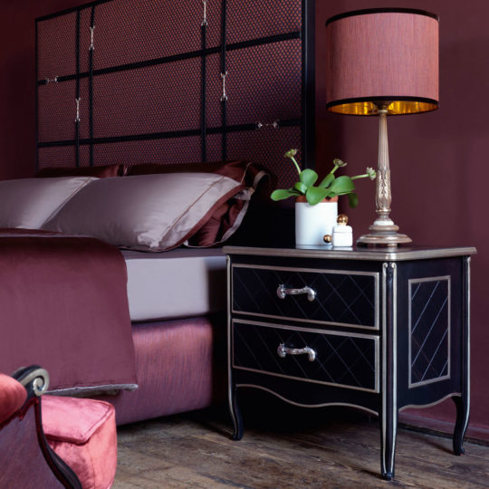 A bedroom showcases a Luxury Italian Horse Handle Bedside Cabinet with two drawers, each accented by silver handles. The cabinet is elegantly topped with a vase containing green plants, a small candle jar, and a gold lamp adorned with a red shade. The bed is dressed in brown and red bedding, complementing the deep red paint on the wall.