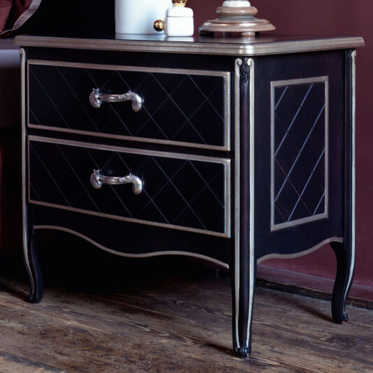 The Luxury Italian Horse Handle Bedside Cabinet showcases a luxurious black finish with two drawers adorned by diamond-patterned fronts and silver horse-shaped handles. This elegant nightstand features curved legs and a polished wooden top, complemented by its placement on a hardwood floor. A lamp with a white base and beige shade adds the finishing touch to this sophisticated setting.