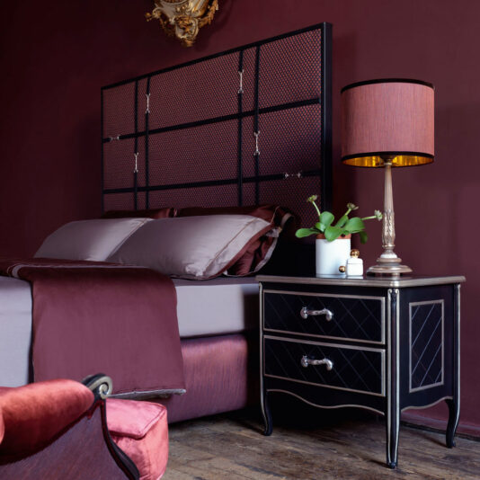 A luxurious bedroom with burgundy walls features a bed with a tall, woven headboard, dressed in gray and burgundy linens. A Luxury Italian Horse Handle Bedside Cabinet holds a stylish lamp with a burgundy shade, a small plant, and other decor items. An upholstered chair is visible in the foreground.