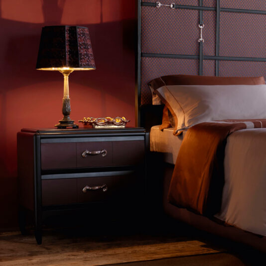 A cozy bedroom features a Luxury Leather Horse Handle Bedside Cabinet with two drawers and a lit table lamp. The background wall is painted in deep red, and the bed is adorned with brown and white bedding. A pair of glasses rests on the Horse Handle nightstand. A soft, warm ambiance fills the room.