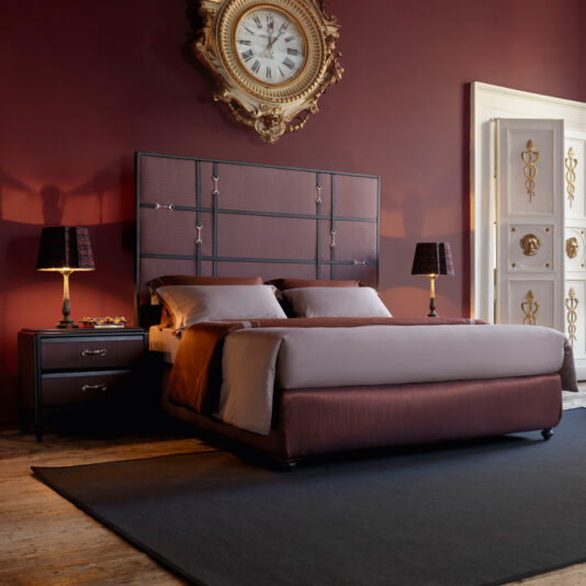 A luxurious bedroom showcasing a spacious bed with burgundy and gray bedding, flanked by two Luxury Leather Horse Handle Bedside Cabinets, each adorned with a stylish lamp. An ornate gold clock is mounted on the rich burgundy wall above the bed. The room features wooden flooring complemented by a dark rug.