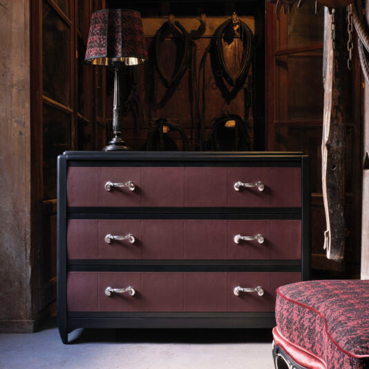 A Luxury Leather Horse Handle Chest Of Drawers made of dark wood with three drawers, each fitted with elegant horse handle accents, sits in a rustic room. A patterned table lamp is on top. In the background, wooden shelves and a wall adorned with old harnesses add charm. A cushioned seat is partially visible on the right.