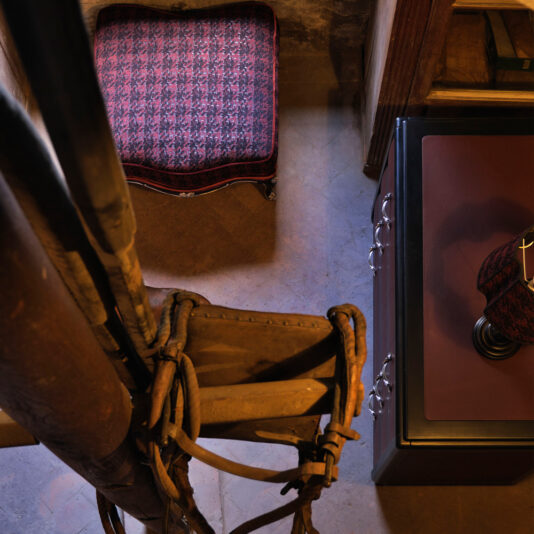 A cozy corner is adorned with a red and black upholstered chair and a table showcasing a matching lamp. A vintage saddle and reins decorate the wall, while the luxurious Luxury Leather Horse Handle Chest Of Drawers adds elegance to the warm, dimly lit ambiance.