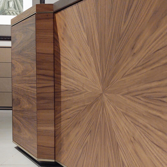A close-up of wooden furniture showcasing a beautiful radial wood grain pattern, with highly detailed texture and natural variations in color. In the background, part of a framed picture and the Luxury Leather Top Desk are partially visible.