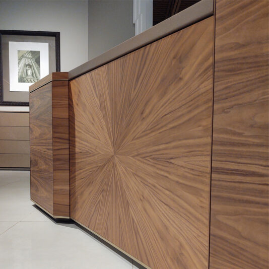 A Luxury Leather Top Desk stands nearby a modern wooden sideboard with a distinctive starburst pattern on the doors in a room with tiled flooring. A framed black and white architectural print hangs on the wall above the stylish sideboard, which features clean lines and a rich wood finish.