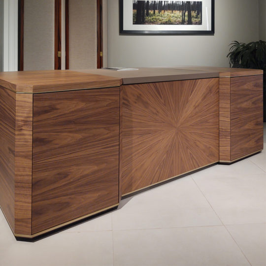 In a minimalist room, the Luxury Leather Top Desk with its sleek, polished finish and distinct geometric patterns on the wooden surface stands prominently. It is positioned on a light-colored tiled floor, with a framed photo of a forest hung on the wall behind it.