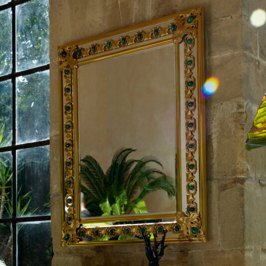 The Luxury Mirror With Green Malachite Details, featuring an ornately-framed design with intricate gold embellishments and green malachite accents, is mounted on a stone wall. It reflects a leafy indoor plant and part of a windowpane, through which light casts a small rainbow pattern on the wall.