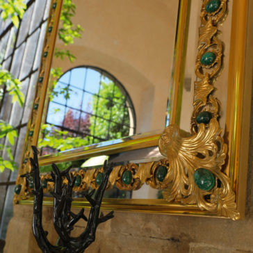 The Luxury Mirror With Green Malachite Details, featuring a gold frame, reflects a window with an arched top. Nearby, a decorative black sculpture resembling tree branches is positioned on a stone ledge while sunlight filters through the surrounding foliage.