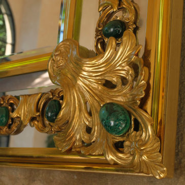 A close-up of the Luxury Mirror With Green Malachite Details showcases its ornate, golden frame adorned with intricately carved designs and gemstones. The elaborate leaf patterns stand out beautifully as the mirror's edge partially reflects sunlight in the background.