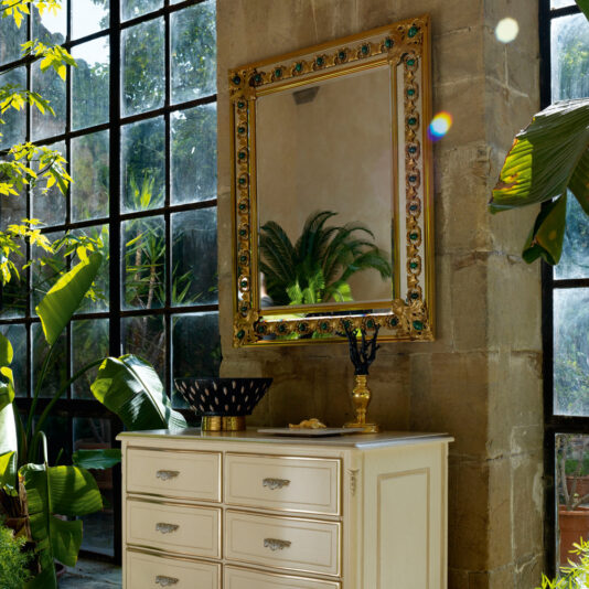 Luxury Mirror With Green Malachite Details A sunlit room with large glass-pane windows showcasing greenery outside. A white dresser with multiple drawers stands against a stone wall, topped with a decorative bowl and ornate candle holder. Above the dresser hangs the Luxury Mirror With Green Malachite Details, reflecting plants.