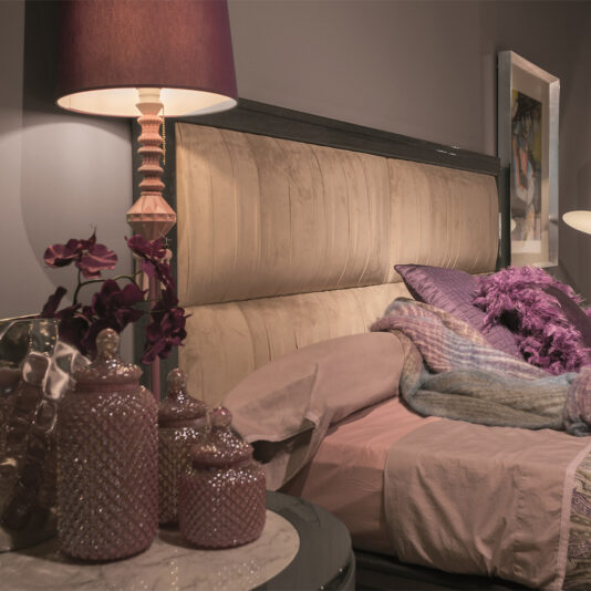 A cozy bedroom features the Luxury Modern Upholstered Bed with beige fabric, adorned with pink and purple bedding. A matching lamp with a pink shade and ceramic jars sit on the bedside table. Purple flowers add a touch of decoration. The room is softly lit, exuding a tranquil ambiance.