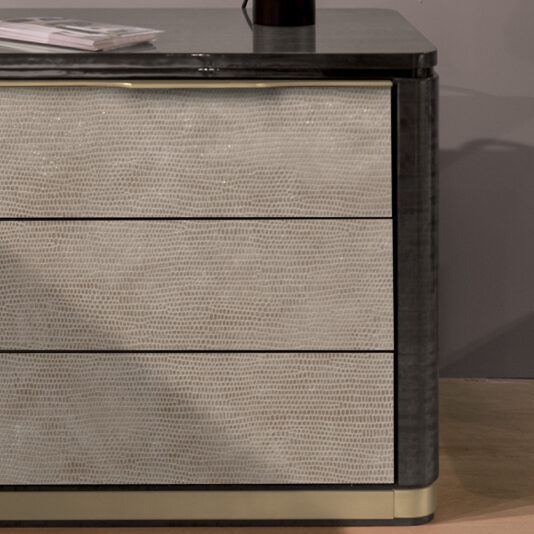 Modern Bedside Table With Leather Front The Modern Bedside Table With Leather Front features a sleek, modern design with a leather front and dark wood frame. It includes three spacious drawers, with a black vase and magazine elegantly displayed on top. The setting is complemented by a muted gray wall in the background and light wooden flooring.