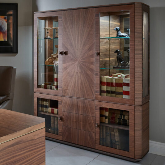 The Luxury Modern Bookcase With Storage, featuring a sunburst wood pattern and glass doors, showcases various items. Inside are ornate figurines of dogs, leather-bound books, and a small sculpture. The cabinet adds elegant charm to the contemporary room.