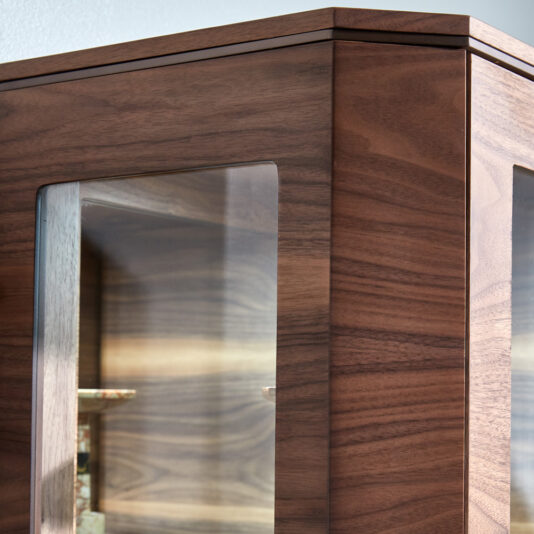 Close-up of the Luxury Modern Bookcase With Storage, featuring glass panel doors. The wood boasts a rich, dark finish, and the glass panels reveal a glimpse of the wooden interior shelves. The cabinet's corner is prominently featured in the image.