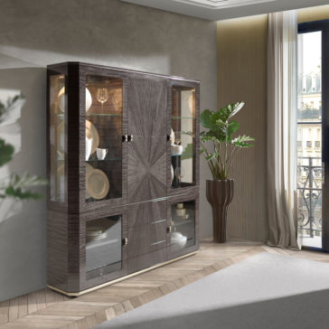A modern living room boasts a Luxury Modern Display Cabinet, complete with glass doors and shelves that beautifully showcase decorative plates and glassware. The room features light-colored walls, wood flooring, a large potted plant, and a window revealing a stunning city view in the background.