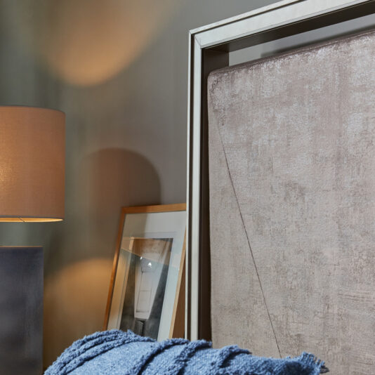 A cozy bedroom corner featuring a soft beige headboard on the Luxury Metal Frame King Size Bed, a modern beige table lamp emitting warm light, a photograph in a wooden frame leaning against the wall, and a navy blue textured blanket. The scene exudes a calm and inviting ambiance.