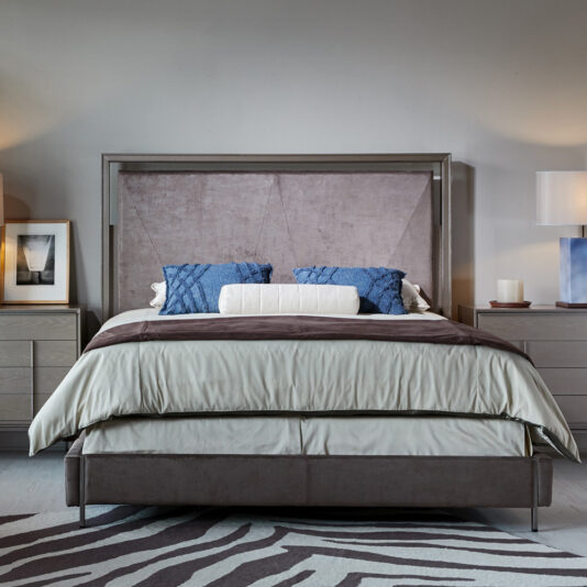 Contemporary bedroom showcasing a Luxury Metal Frame King Size Bed adorned with a spacious, padded headboard. The bed is dressed in beige and white linens, complemented by two blue decorative pillows. Flanking the bed are matching nightstands each topped with lamps and framed artwork, while a zebra-striped rug adorns the floor.
