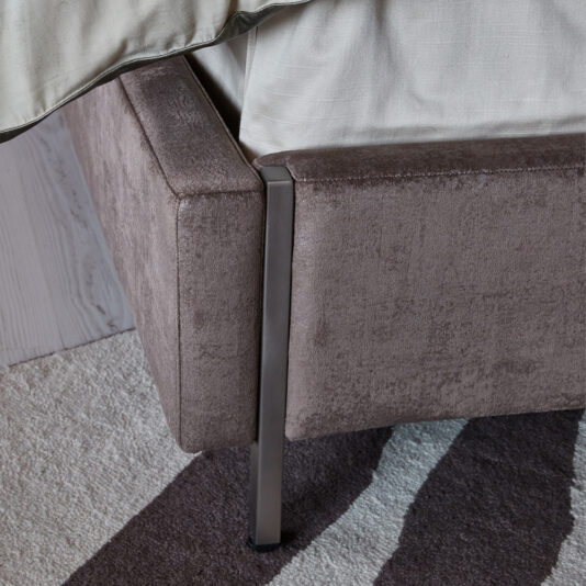 A close-up view of the Luxury Metal Frame King Size Bed corner, upholstered in soft brown fabric. The sleek metal legs beautifully complement the light beige bedspread. The bed is placed on a rug with a brown and white pattern, enhancing its elegant appeal.