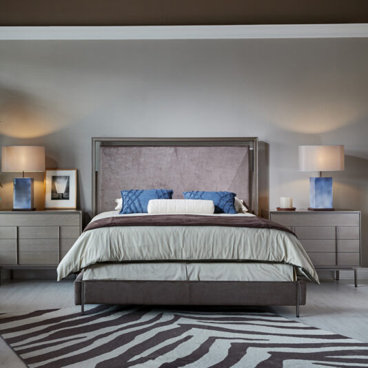 A modern bedroom featuring a Luxury Metal Frame King Size Bed adorned with light gray bedding and two blue pillows. The bed is flanked by two wooden nightstands with drawers, each topped with a blue and white lamp, a framed picture, and some books. A zebra-striped rug lies under the bed.