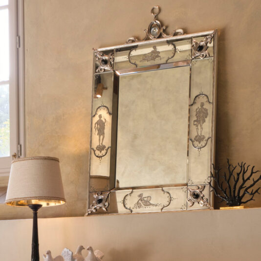 A Luxury Venetian Mirror With Black Onyx Details, showcasing a rich vintage-style frame adorned with etched designs of classical figures and floral motifs, hangs elegantly on a beige wall near a window. It is beautifully complemented by a table lamp and an artistic small sculpture of a tree branch nearby.