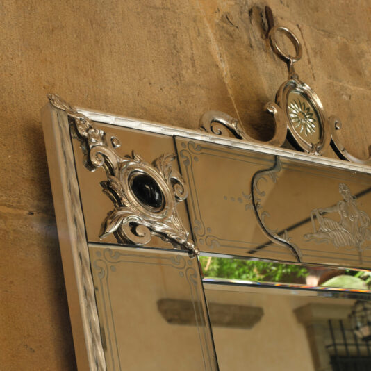 A close-up view of the Luxury Venetian Mirror With Black Onyx Details, showcasing its intricate silver craftsmanship and black onyx embellishments. The mirror is mounted on a textured stone wall, reflecting an outdoor scene with blurred greenery and architectural elements in the background.