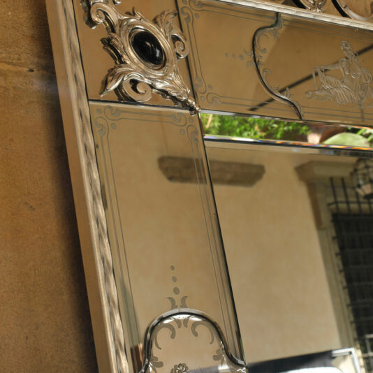 A close-up of the Luxury Venetian Mirror With Black Onyx Details reveals its intricate carvings and an emblem featuring black onyx accents. This mirror also boasts etched designs, reflecting a section of a courtyard with a stone wall and lush greenery. The overall aesthetic is both ornate and classical.