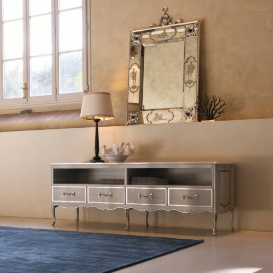 A stylish room corner showcases a wooden dresser with a metallic finish, adorned with decorative bowls and a table lamp. Mounted above is the Luxury Venetian Mirror With Black Onyx Details on a textured beige wall. A blue rug graces the floor, and natural light streams in through a nearby window.