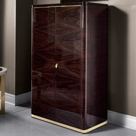 The Modern Armoire With Brass Accents is a tall, glossy wooden cabinet featuring a polished brown finish and brass detailing at the base and top. This contemporary piece boasts two front doors, adorned with a small gold handle in the center, and is placed against a beige wall on a light gray tiled floor.
