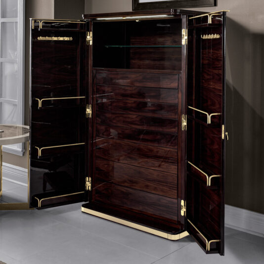 The Modern Armoire With Brass Accents, featuring a sleek dark brown wooden construction with a glossy finish, stands open. This contemporary piece showcases a glass shelf at the top, multiple wooden shelves below, and doors equipped with built-in storage racks. It is elegantly positioned against a wall in a modern room.