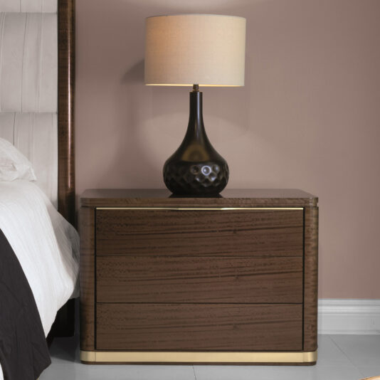The **Modern Bedside Table With Brass Trim** with three drawers stands beside a bed. A black table lamp with a beige lampshade is positioned on top, providing a soft light. The wall behind is light brown, and part of the bed with white and dark bedding is visible.