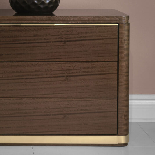 Modern Bedside Table With Brass Trim A modern wooden dresser with three smooth drawers, accented by brass trim at the top and bottom, stands against a beige wall. The dresser has a dark, polished finish, and a black textured vase sits on top—perfectly complementing your Modern Bedside Table With Brass Trim.