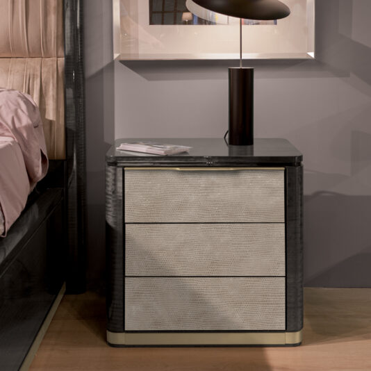 A modern bedside table with a dark marble top and three light-colored drawers with a woven texture. A black lamp sits on the table next to a small stack of papers. The background features a framed picture on a gray wall and part of a bed with beige bedding.