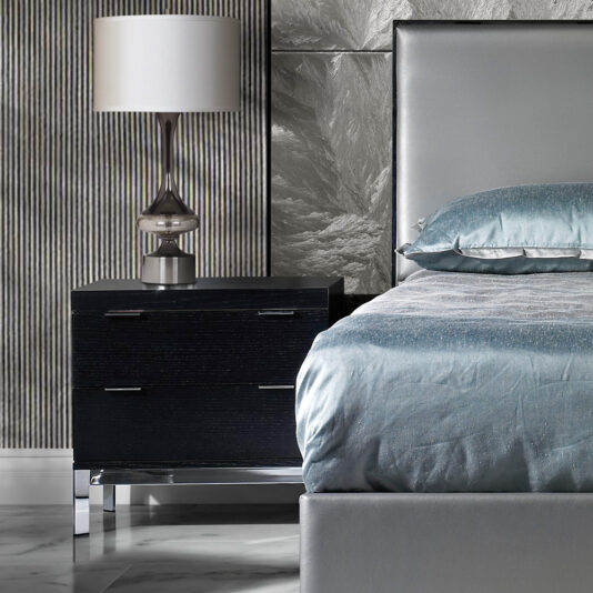 A modern bedroom corner features a gray upholstered bed adorned with blue bedding. A sleek, contemporary Modern Black Bedside Table with a chrome base holds a sophisticated silver lamp with a white shade. The background showcases a textured gray wall and an eye-catching ribbed design panel.