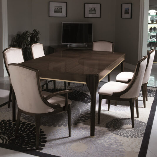 A dining room showcasing the Modern Maple Extendable Dining Table, encircled by six upholstered chairs. Beneath the table lies a patterned rug, while a TV sits on a cabinet against the back wall. The walls are adorned with framed pictures, and potted plants enhance the corners of the room.