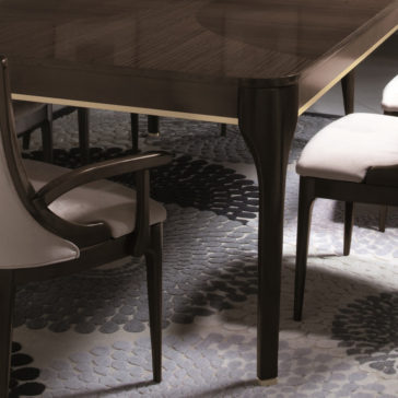 Modern Maple Extendable Dining Table A close-up view of the Modern Maple Extendable Dining Table with cushioned chairs featuring light upholstery. The table surface reflects light, and the gray patterned rug beneath adds texture to the scene. The setting appears elegant and contemporary.