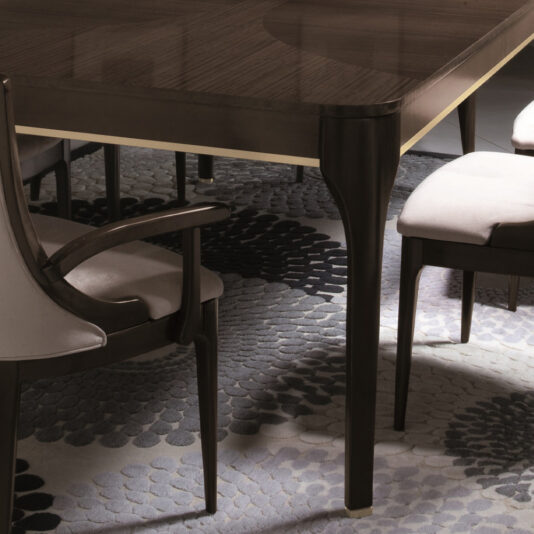 Modern Maple Extendable Dining Table A close-up view of the Modern Maple Extendable Dining Table with cushioned chairs featuring light upholstery. The table surface reflects light, and the gray patterned rug beneath adds texture to the scene. The setting appears elegant and contemporary.