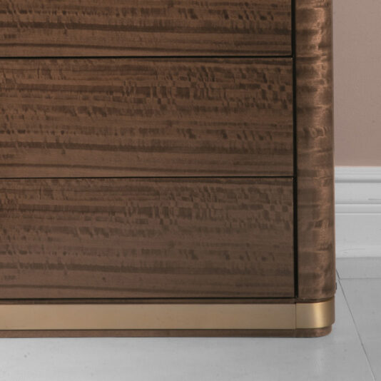 A close-up image of the Modern Tallboy Chest Of Drawers, showcasing its rich, dark wood finish. The chest features a slightly textured surface with three visible horizontal drawers. Its right edge is rounded, and it stands gracefully on a light-colored floor.