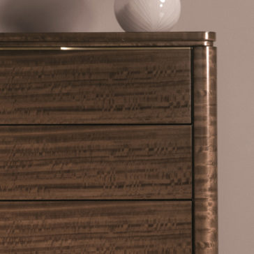 Close-up of the Modern Tallboy Chest Of Drawers featuring a rich, wavy grain pattern and showcasing three drawers. The chest boasts a smooth, polished finish with rounded edges. On top sits a white decorative vase with a geometric design, set against a plain wall backdrop.