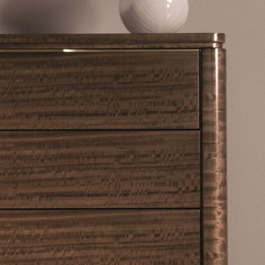 Close-up of the Modern Tallboy Chest Of Drawers featuring a rich, wavy grain pattern and showcasing three drawers. The chest boasts a smooth, polished finish with rounded edges. On top sits a white decorative vase with a geometric design, set against a plain wall backdrop.