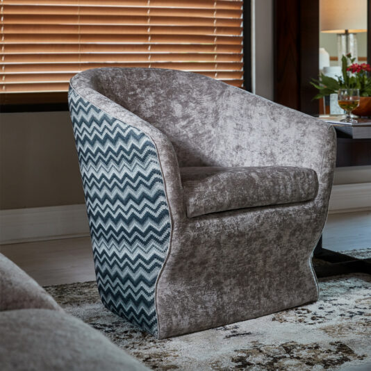 A modern, upholstered armchair featuring a gray, textured fabric on the front and a blue and white zigzag pattern on the back and sides. It is placed in a contemporary living room with a window covered by horizontal blinds, revealing some light.