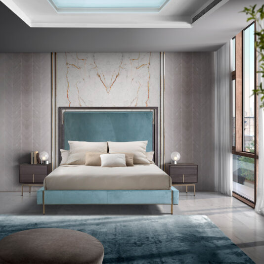 A contemporary bedroom showcases a spacious bed adorned with a teal upholstered headboard and understated bedding. The room is enhanced with Luxury Leather Horse Handle Bedside Cabinets, each graced with horse handle accents and accompanied by round lamps. A marble accent wall, chevron-patterned walls, floor-to-ceiling windows, and a soft blue rug add to the room's sophisticated ambiance.