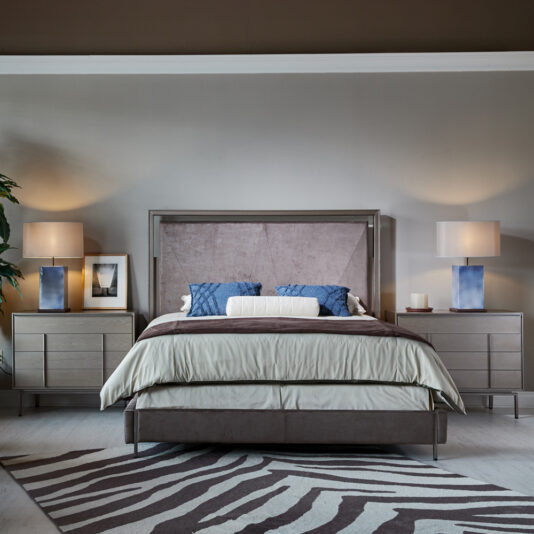 Modern Elegant Chest Of Drawers A neatly arranged bedroom showcases a large bed adorned with neutral bedding and blue accent pillows. Nightstands on either side of the bed hold blue and white lamps. The gray walls provide a calming backdrop, complemented by indoor plants and framed pictures for added decor. On the floor, near the Modern Elegant Chest Of Drawers, lays a striking zebra-patterned rug.
