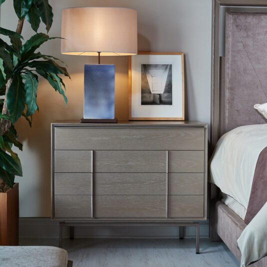 A modern, elegant bedroom showcases the Modern Elegant Chest Of Drawers, featuring a light wooden finish and sleek, minimalist design. On the dresser sits a rectangular lamp with a beige shade and a framed black-and-white photograph. A bed with a gray headboard is partially visible to the right, next to a large green plant.
