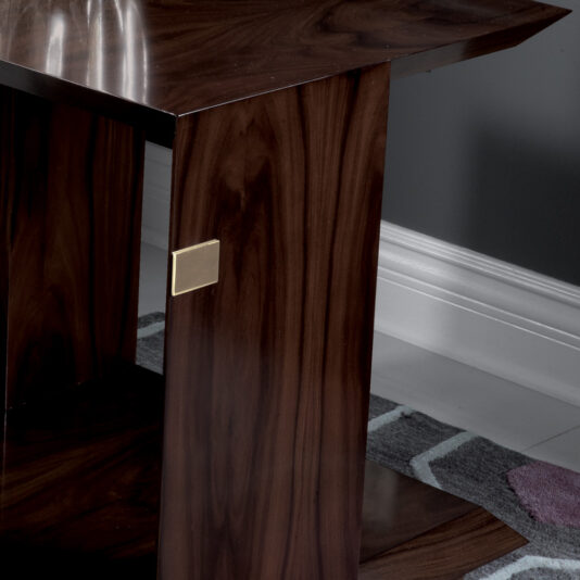 The Modern Elegant Side Table, featuring a dark wooden finish and a small rectangular metallic inlay on one leg, stands gracefully on a grey and white patterned rug. It is situated adjacent to a white baseboard and grey wall.