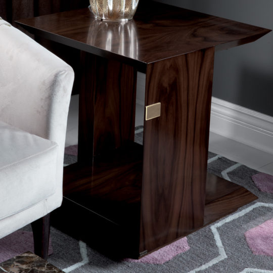 A Modern Elegant Side Table made of dark wood, equipped with a lower shelf, is positioned next to a light-colored upholstered sofa. The table features a sleek design with clean lines and includes a small golden accent on one side. It rests on a patterned rug in shades of grey and pink.