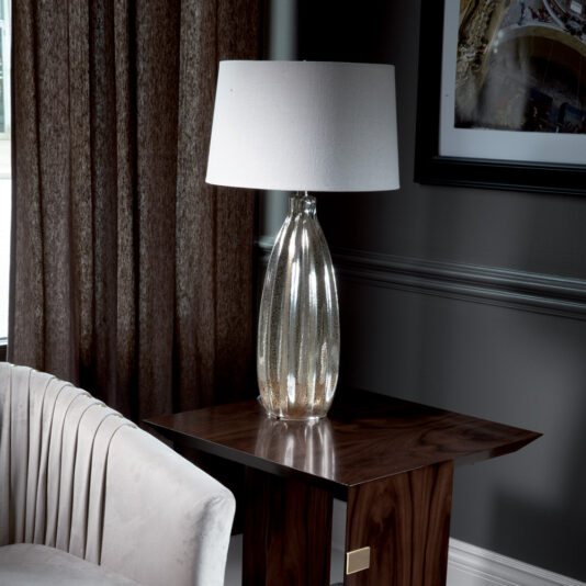 A silver metallic table lamp with a white shade sits on a Modern Elegant Side Table made of dark wood. To the left is a beige upholstered chair, and behind the setup is a window with brown curtains and part of a framed picture hanging on a dark gray wall.