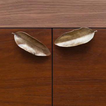 Modern Elegant Sideboard A close-up of two cabinet doors with wooden surfaces featuring handles shaped like metallic leaves. The leaves are oriented in opposite directions and have a smooth, reflective finish that contrasts with the wood grain of the cabinets.