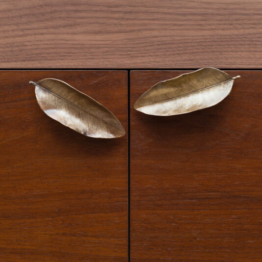 A close-up of two cabinet doors with wooden surfaces featuring handles shaped like metallic leaves. The leaves are oriented in opposite directions and have a smooth, reflective finish that contrasts with the wood grain of the cabinets.