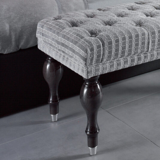 Modern Grey Button Upholstered Bench A close-up image showcases the Modern Grey Button Upholstered Bench, which boasts button-tufted upholstery in a checkered pattern and dark wooden legs with metallic caps at the bottom. The bench is placed on a smooth, gray floor near the edge of a bed.