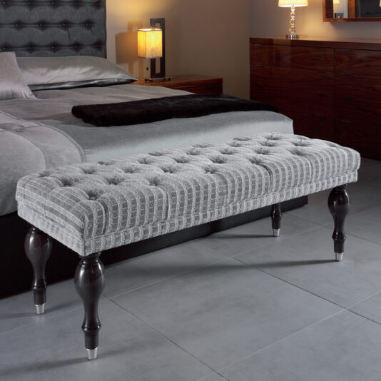 In a luxurious bedroom, the Modern Grey Button Upholstered Bench graces the foot of the bed. The bed is adorned with a tufted grey headboard, coordinated grey bedding, and an elegant black throw blanket. Wooden nightstands with lamps and a dark wooden dresser paired with a mirror enhance the chic ambiance.